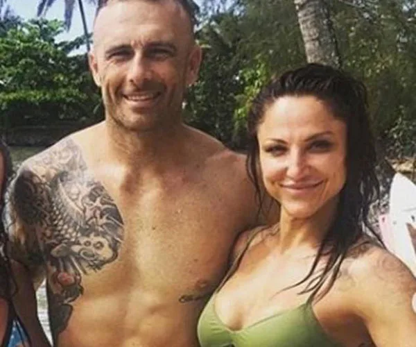 A shirtless man with a tattooed arm and a woman in a bikini smile on a beach with tropical trees in the background.