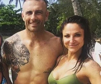 A shirtless man with a tattooed arm and a woman in a bikini smile on a beach with tropical trees in the background.