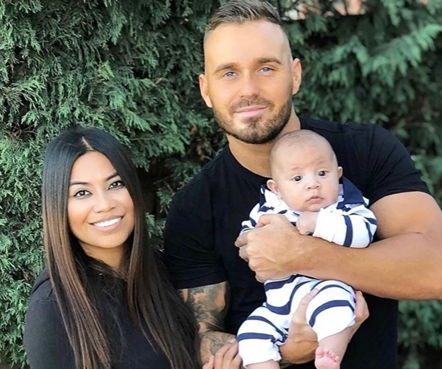 Family posing together outdoors, a woman and man smiling, holding a baby in striped clothes.