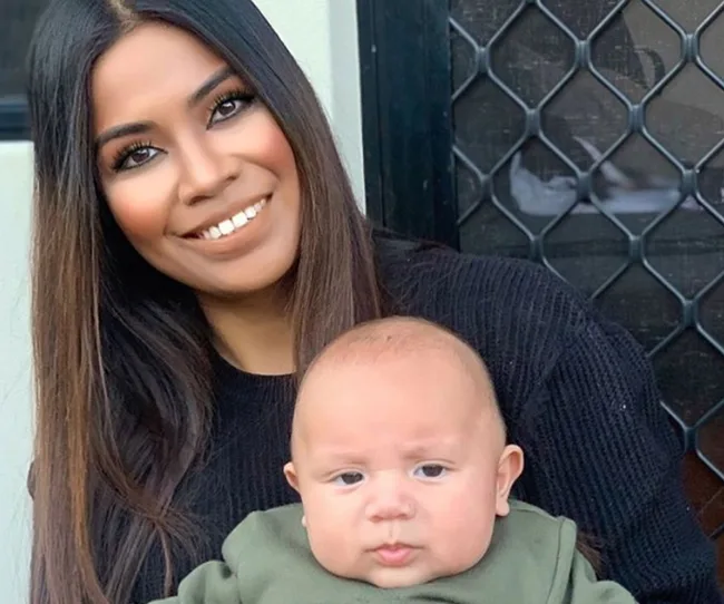 A woman with long hair smiles while holding a baby wearing a green outfit in front of a screened door.