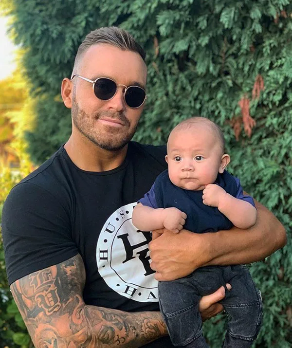 Man in sunglasses holds a baby outdoors with greenery in the background.