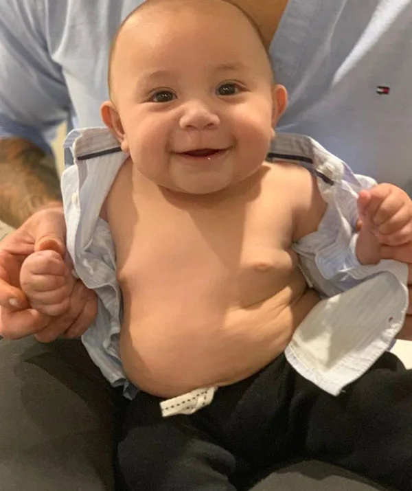 Smiling baby wearing an open shirt and sitting on someone's lap.