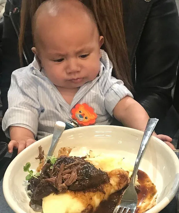 Baby looking confusedly at a plate of mashed potatoes, gravy, and meat while seated on an adult's lap.