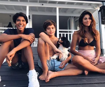 Three people seated on a porch, smiling; two boys and a woman with a dog. Casual outdoor setting.
