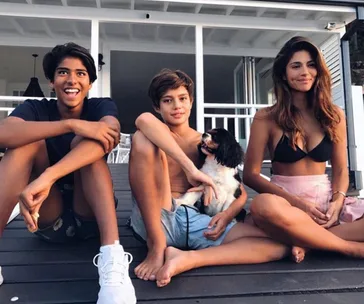 Three people seated on a porch, smiling; two boys and a woman with a dog. Casual outdoor setting.