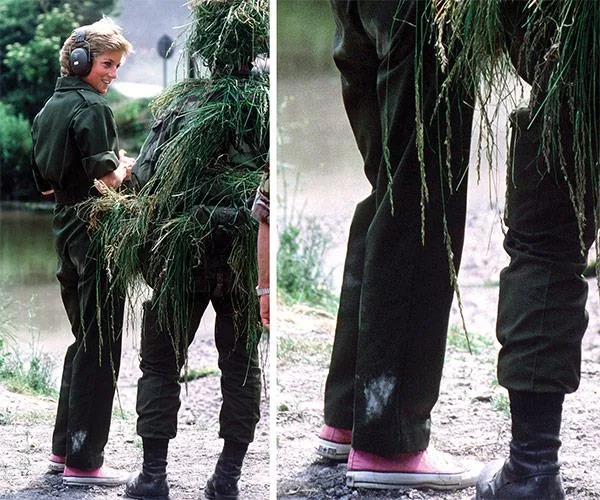 Woman in a military outfit with pink sneakers stands next to a camouflaged person by a pond.