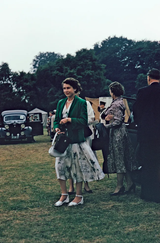 A woman in a green jacket and floral dress walks on grass, with vintage cars and trees in the background.