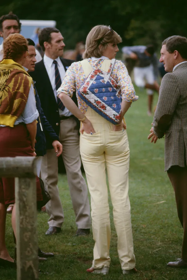 A woman in yellow overalls and a floral shirt stands in a group outdoors, with a floral quilted bag on her back.