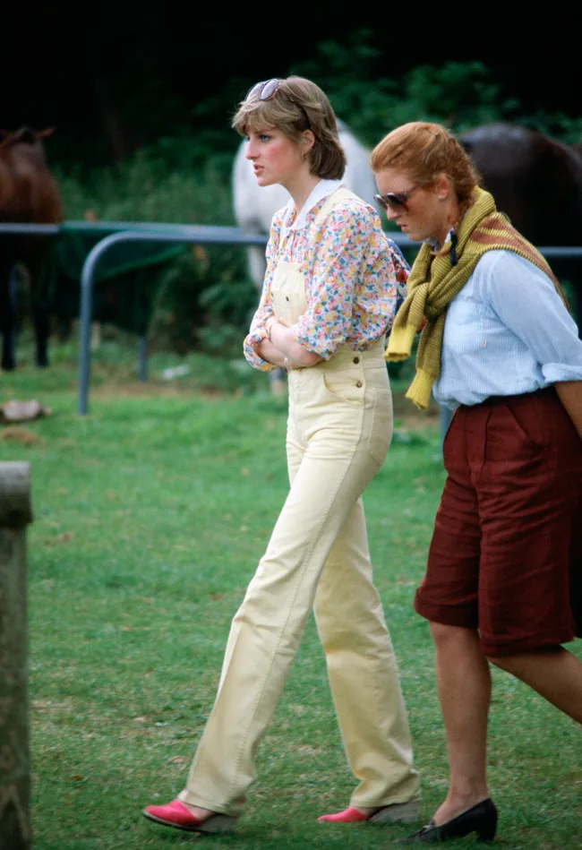 Two women walking on grass, one in yellow overalls with sunglasses, the other in red shorts and a sweater draped over shoulders.