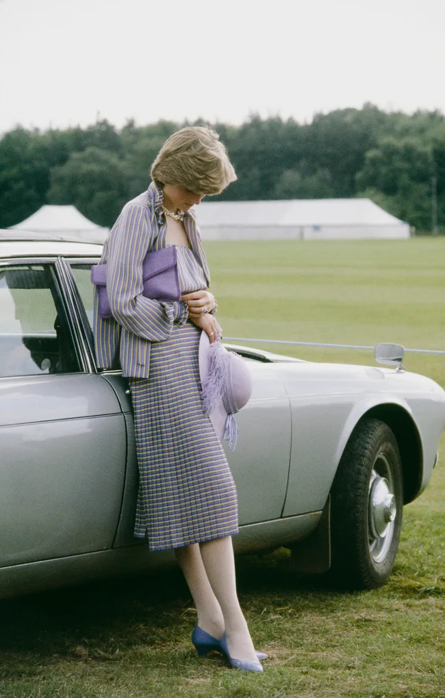 A woman in a striped outfit leans against a car, holding a purple clutch and hat, with a green field in the background.