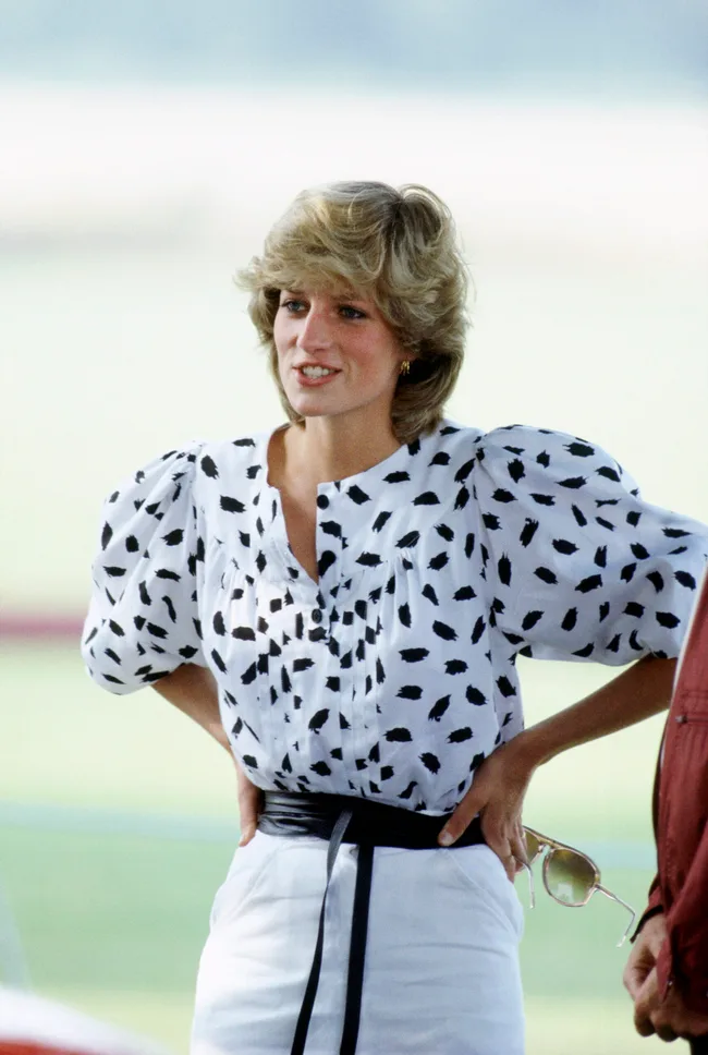 A woman in a white and black polka dot blouse with hands on her waist, holding sunglasses, smiling outdoors.