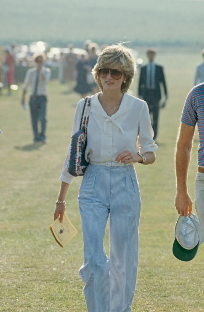 A woman in sunglasses and a white blouse walks outdoors, holding a paper and patterned bag, with people in the background.