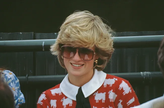 A person with short blonde hair wearing sunglasses and a red sweater with white sheep pattern, smiling.