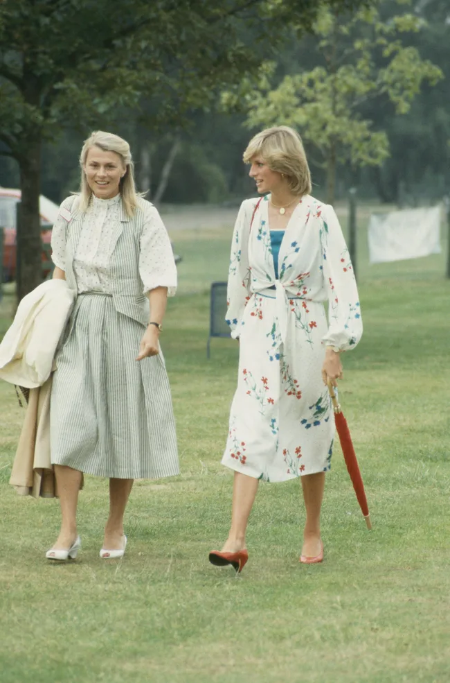 Two women walking on grass, one in a striped dress holding a coat, the other in a floral dress holding a red umbrella.