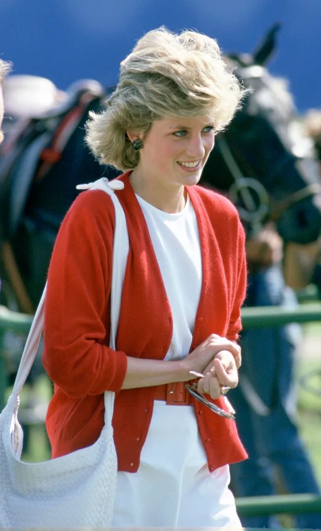 A woman in a red cardigan and white outfit, smiling outdoors, with a white bag and sunglasses in her hand.
