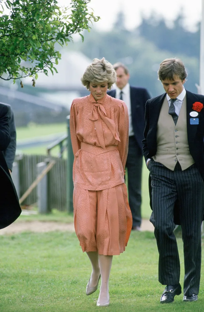 A woman in a peach dress walks on grass with a man in a suit, both looking down, with trees and blurred background.