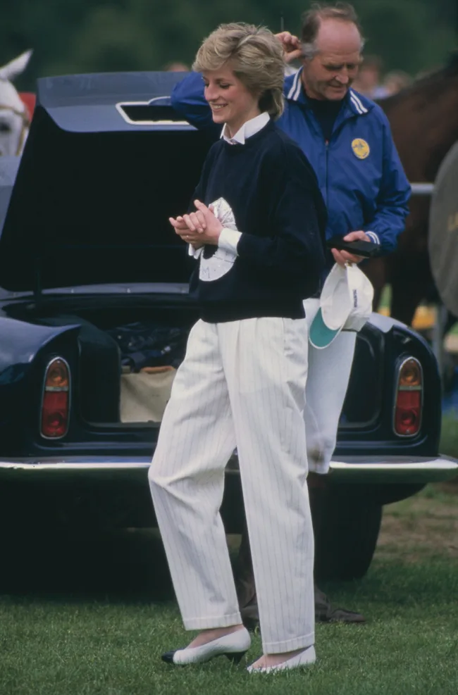 A woman smiling in a black sweater and white pants, standing by an open car trunk on grass, with a man nearby.