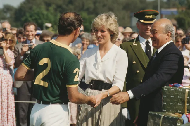 A woman in white shakes hands with a man in a green polo shirt labeled "2" at an outdoor event, surrounded by a crowd.