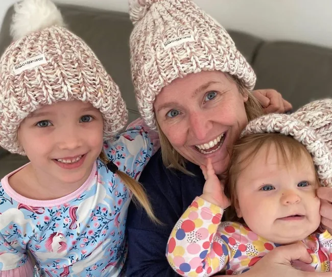 Mother and two children in matching knitted hats, smiling together indoors.