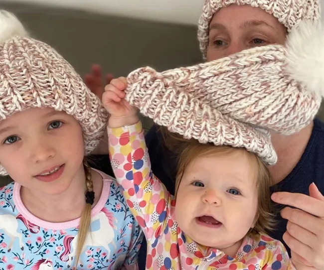 Three people wearing matching knit hats, a child holding an adult's hat down, smiling, indoors.