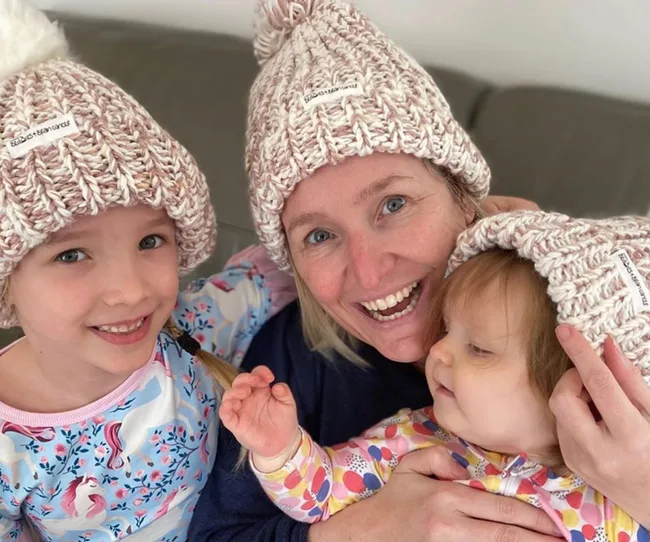 A smiling woman and two young children, all wearing matching knit hats, sitting together indoors.