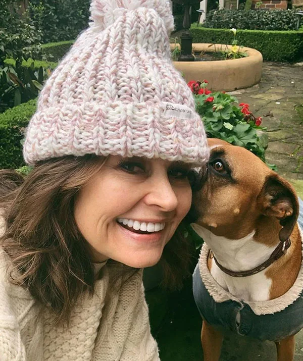 Woman in knit hat smiles while a dog nuzzles her cheek in a garden setting.
