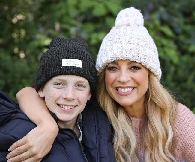 Two people wearing knit hats, smiling outdoors with greenery in the background.