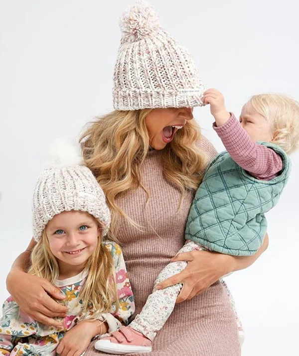 Mom and two kids in cozy hats, smiling and playing together indoors.