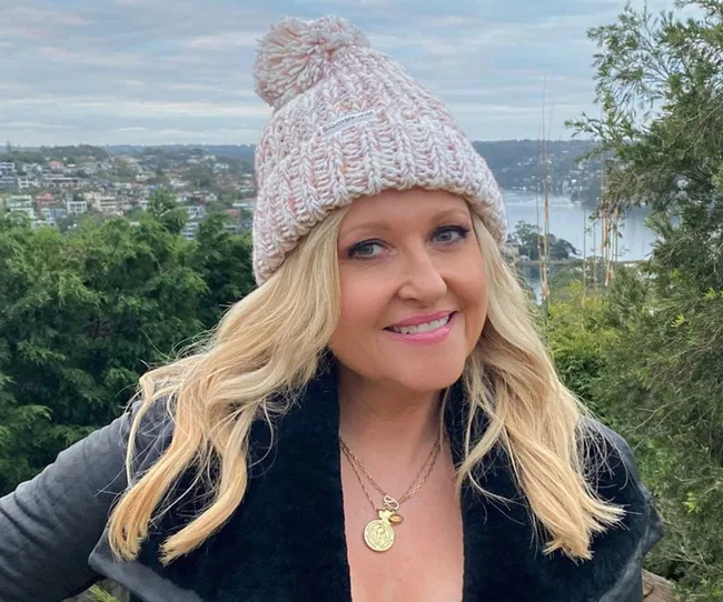 Blonde woman in a knit hat smiles outdoors with a scenic view in the background.