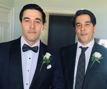 Two formally dressed men with matching rose boutonnieres smiling at the camera.