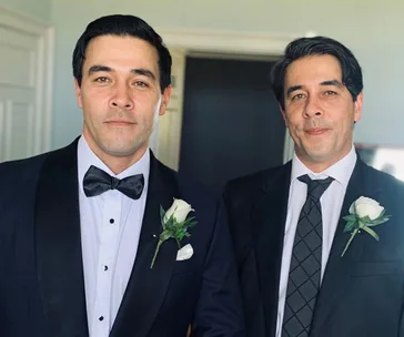 Two formally dressed men with matching rose boutonnieres smiling at the camera.