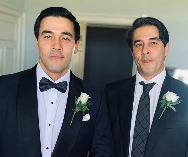 Two formally dressed men with matching rose boutonnieres smiling at the camera.