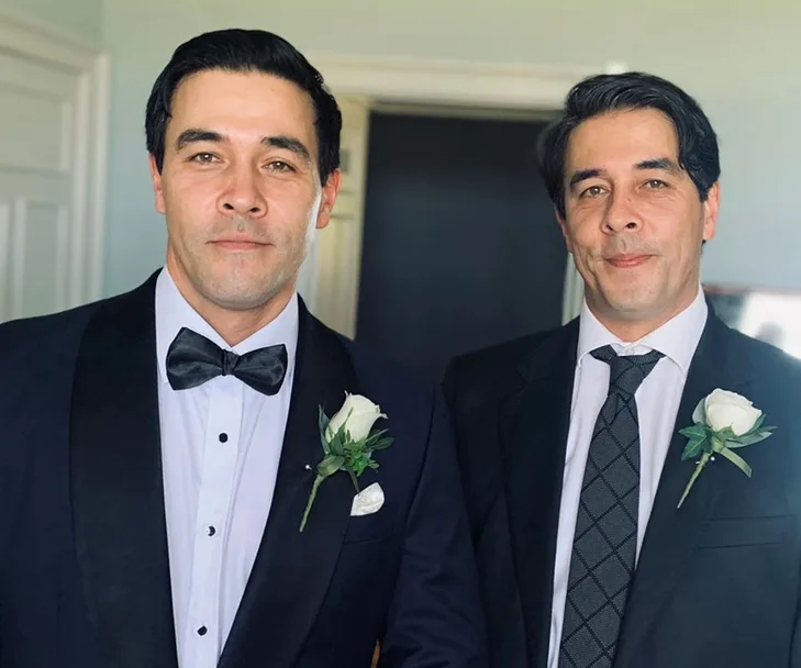 Two formally dressed men with matching rose boutonnieres smiling at the camera.