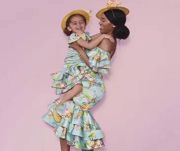 A woman and girl in matching floral dresses and hats smile joyfully against a pink background.