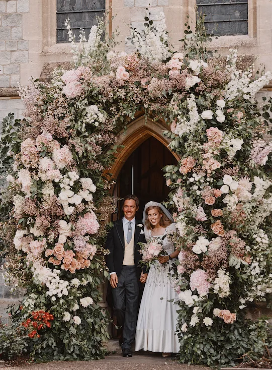 Bride and groom under a large floral arch at a church entrance.
