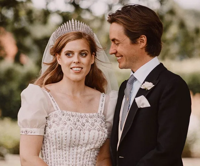 A bride in a beaded gown and diamond tiara stands beside a groom in a suit, both smiling outdoors.