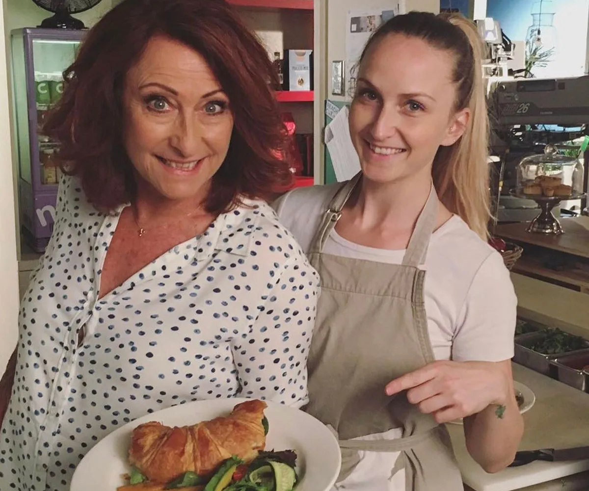 Two women smiling in a kitchen, one holding a plate with a croissant and salad, the other pointing at the dish.