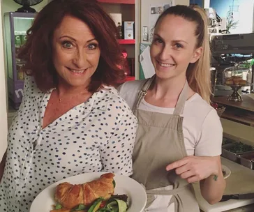 Two women smiling in a kitchen, one holding a plate with a croissant and salad, the other pointing at the dish.