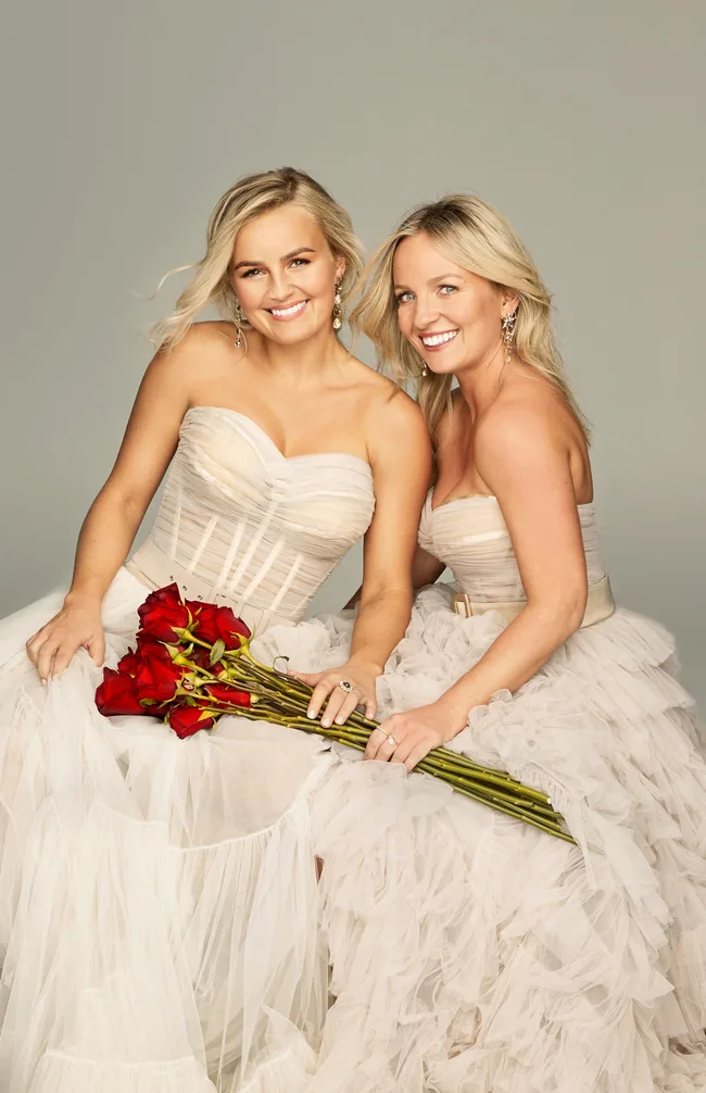 Two women in matching strapless gowns holding a bouquet of red roses, smiling at the camera.
