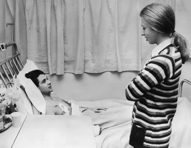 A woman in a striped sweater visits a man in a hospital bed, engaging in conversation.