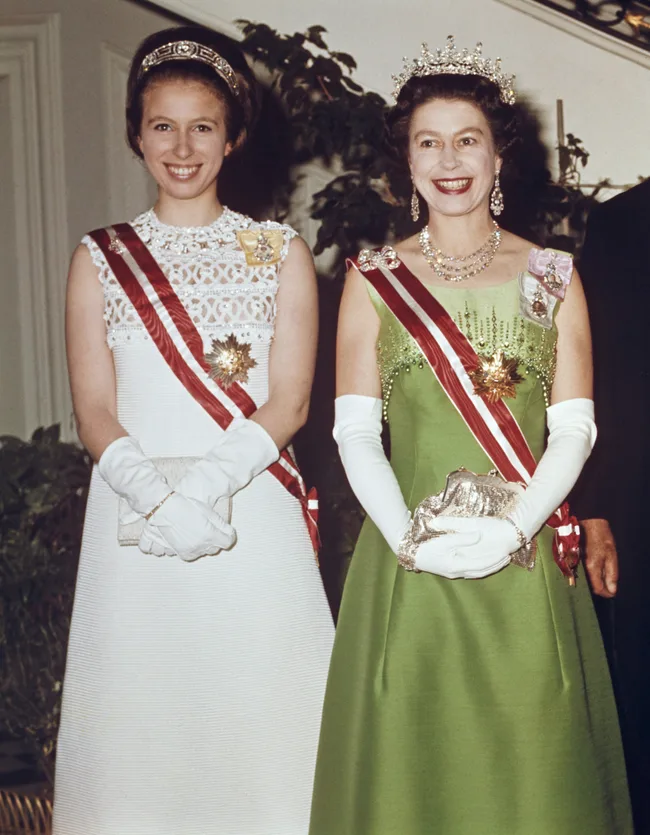 Two women in formal dresses with tiaras and sashes, smiling at a formal event.