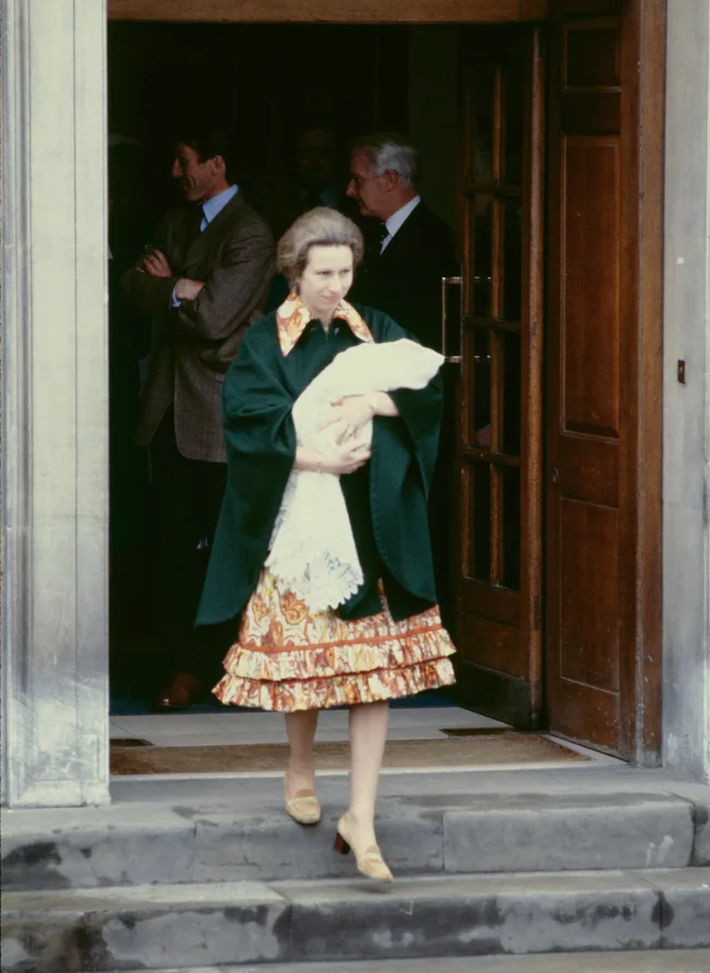 Woman in floral dress and green cape holding a baby, walking down steps from a building with people in the background.