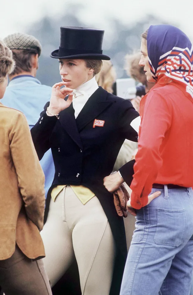 Equestrian in formal attire, top hat, and Union Jack badge, talking with others outdoors.