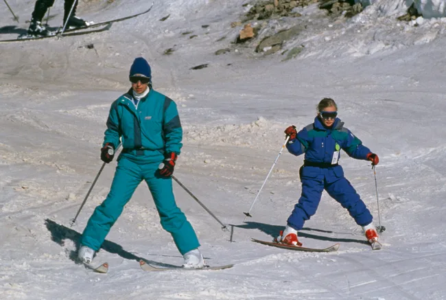 Two people skiing on snowy slopes, wearing teal and blue ski suits and goggles.