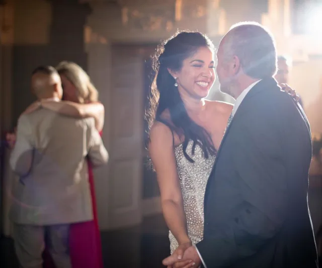 A woman in a white dress smiles while dancing with an older man at a formal event, another couple is dancing in the background.
