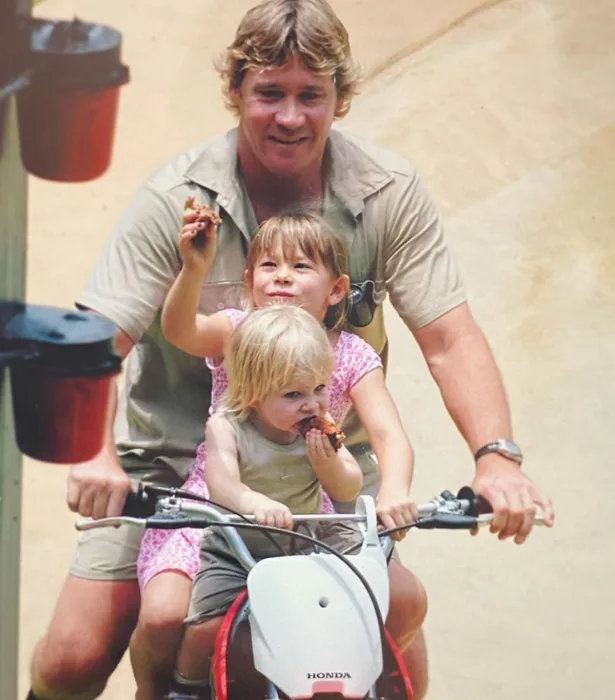 Man riding a small Honda bike with two young children eating snacks, joyfully seated on his lap.