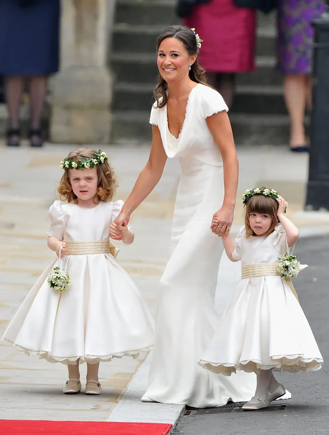 Female in white dress with two young girls in matching white dresses and flower crowns on a red carpet.