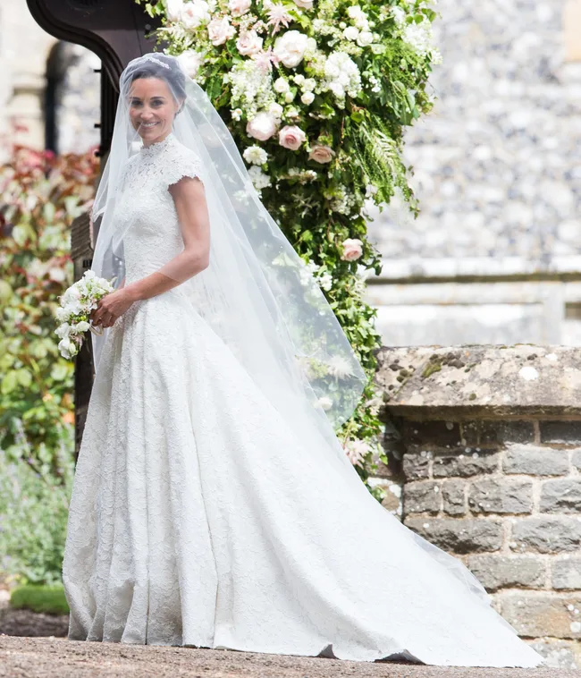 A bride in a lace gown and veil stands outside, holding a bouquet, with floral arch in the background.