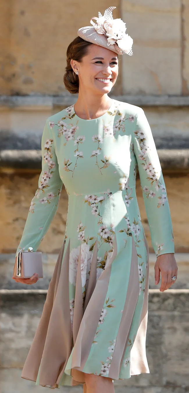 A woman in a floral dress and hat, smiling, holding a clutch at an outdoor event.