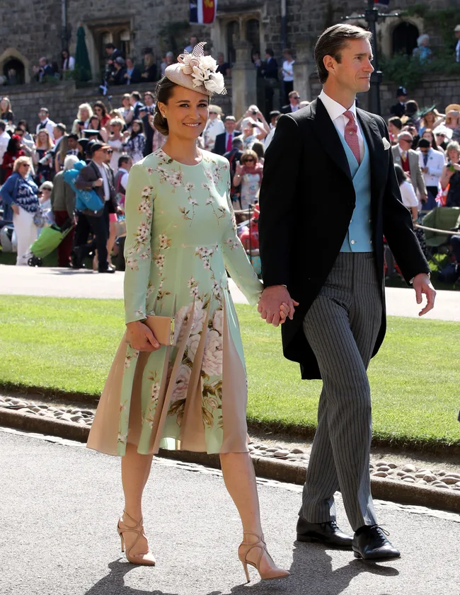 A couple in formal attire holding hands, with the woman in a floral dress and fascinator, amidst a crowd outside a stone building.